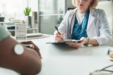 Middle aged Caucasian female doctor consulting Black adult patient with diabetes, writing notes on clipboard while patient with glucose monitor on arm sitting across medical office desk
