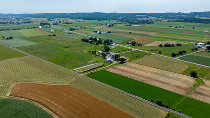 A vast, open landscape showcases fields of varying colors and textures, with a few farmhouses scattered throughout. The clear sky allows for bright sunlight illuminating the fields.