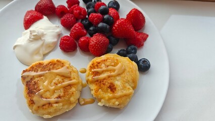 Delicious syrniki with caramel topping served with fresh berries and sour cream on a white plate. Concept of homemade breakfast, healthy eating, indulgence and morning comfort food