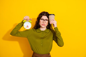Young female holding clock and coffee cup, frustrated late morning concept on vibrant yellow...
