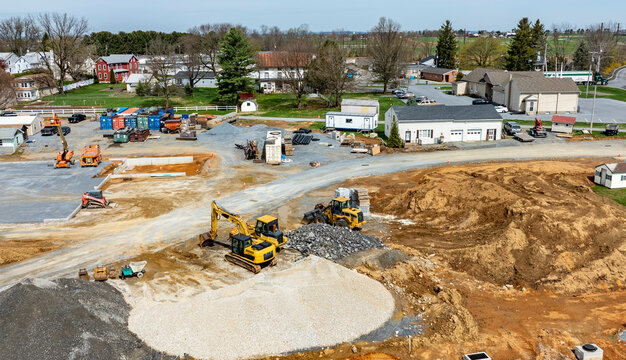 Heavy machinery operates on a large construction site, with earth being moved and materials arranged. Nearby buildings and green fields provide a backdrop to the ongoing development.