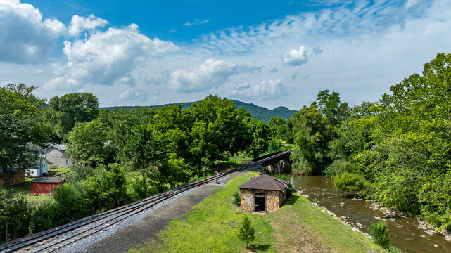 A tranquil view showcases railway tracks running along a winding creek, surrounded by dense trees and rolling hills.