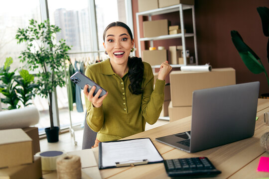 Cheerful businesswoman celebrates success while working remotely in her modern fashion atelier in a stylish city studio