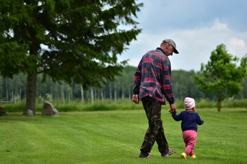 Senior man and toddler girl walking hand in hand on a green lawn in the countryside. Rear view of...