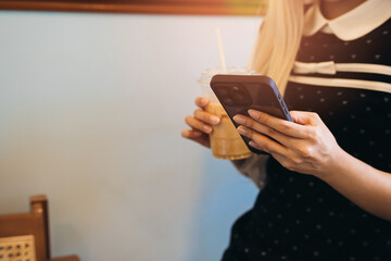 Woman using smartphone. Young woman with long hair holding iced coffee and smartphone in a cozy cafe setting, enjoying a moment of relaxation and connection