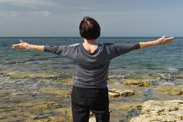  woman standing with artrits arms outstretched on a rocky beach by the calm sea, concept of wellness, relaxation, outdoor tourism