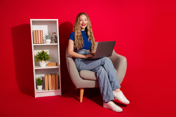 Young woman with long blonde hair working on a laptop sitting on a comfortable armchair against a...