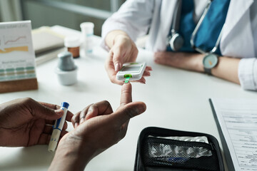 Black man having blood glucose level checked by female doctor in medical office, doctor holding glucometer while patient pricking finger, diabetes management consultation scene