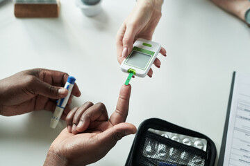 Black adult hand receiving blood glucose test from Caucasian female medical professional in clinic,...