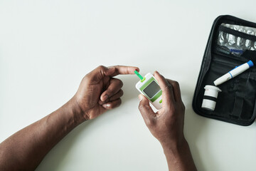 Black man using blood glucose meter checking blood sugar level with test strip on finger in medical office, diabetes management concept, diabetes supplies and glucometer visible on table