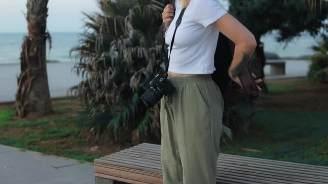 Young photographer preparing camera and backpack while sitting on park bench near seaside, readying equipment for upcoming photo expedition with professional concentration and creative anticipation
