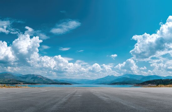 Empty asphalt road stretches to a serene lake and mountains under a vast blue sky with fluffy white clouds