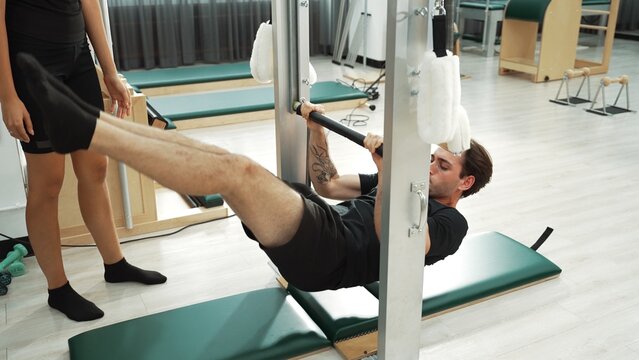 Professional pilates instructor guiding a client during an inversion stretch on a Pilates frame at gym. Man hanging on bar for improving core stability and spinal alignment. Wellbeing. Habituate.