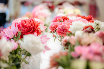 Rows of vibrant peony flowers in various shades of pink, red, and white are displayed in vases on tables at an indoor floral exhibition.