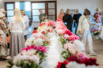 Rows of colorful peonies are displayed on long tables at a flower exhibition, with people in the background admiring the floral arrangements.