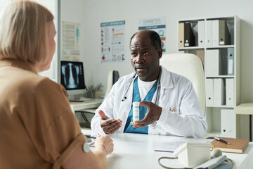 Obraz premium Middle aged Black male doctor explaining medication to middle aged Caucasian woman patient during diabetes consultation in medical office, prescription bottle visible on desk