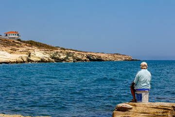 Senior Man Fishing on Rocky Mediterranean Coast in Cyprus, Elderly man sits fishing on rocky shore, overlooking the calm blue sea and coastal cliffs.
