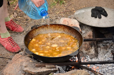 Outdoor cooking process with water being poured into a cauldron of bubbling stew over a campfire....
