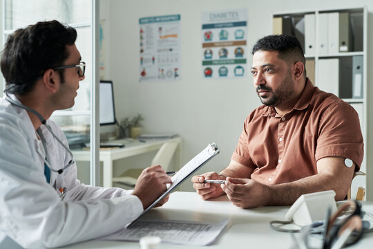 Middle aged Caucasian man with diabetes consulting male doctor in medical office, patient holding glucose meter while discussing health management, medical posters visible in background