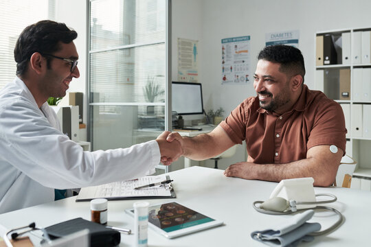 Middle aged Caucasian man with diabetes wearing glucose monitor sitting at desk shaking hands with young adult Caucasian male doctor during medical consultation in clinic