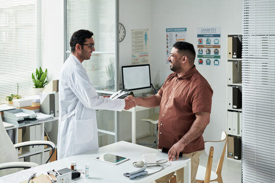 Middle aged Caucasian man visiting doctor in medical office, shaking hands with young adult South Asian male physician, discussing diabetes management with medical equipment on desk