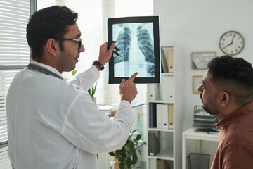 Middle aged Caucasian male doctor showing chest X-ray to middle aged Black man patient during...