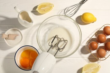 Hand mixer, egg whites in bowl, eggs and other products on white wooden table, flat lay