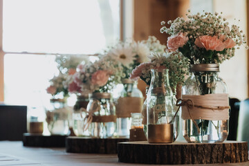 Row of mason jar vases on wood slice slabs filled with pink carnations, white daisies and baby’s breath on a rustic reception table