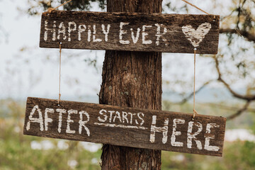 Rustic wooden hanging wedding sign on a tree trunk with white painted text “Happily Ever After Starts Here” in an outdoor woodland setting