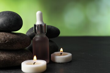 Spa composition with stones, dropper and burning candles on dark table against blurred green background, closeup. Space for text