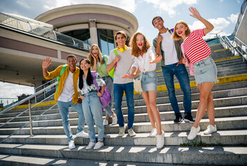 Group of cheerful young friends enjoying sunny day standing outdoors on city stairs, symbolizing...