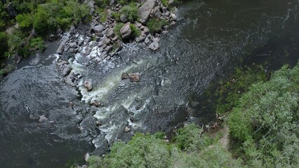 Aerial view of rocks across a section of a flowing stream. Trees and large stone boulders around. Gusts of wind swaying tree branches.