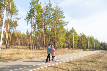 Little girl riding scooter while walking with her grandmother in the forest on a sunny day