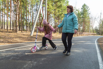 Little girl riding scooter while walking with her grandmother in the forest on a sunny day
