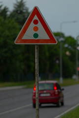 A bright red car is driving swiftly past a triangular traffic sign that is positioned along the roadway, catching the attention of onlookers