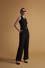 Elegant woman in a sleeveless black vest and wide-leg trousers posing with sunglasses against a beige background. Stylish, powerful, modern fashion concept.