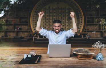 Male freelancer expressing excitement and success with laptop in modern interior, showcasing strong...