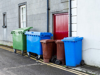 A row of trash cans and bins are lined up on the sidewalk by and old house. All plastic containers...