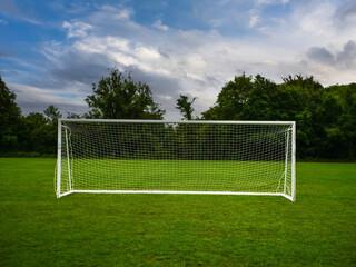 Soccer or football goal posts on clean green grass in foreground, dark trees and dramatic cloudy sky in the background. Popular sport equipment and training ground. Professional club pitch.