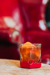 Amber cocktail in red-wax-dipped rock glass with spherical ice and curled orange-peel garnish on wooden table before red leather armchair