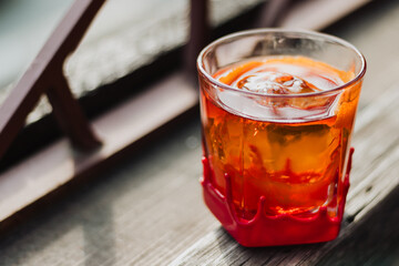Red-wax-dipped rock glass with amber cocktail, spherical ice, and curled orange-peel garnish on weathered balcony railing overlooking waterfront in soft natural light