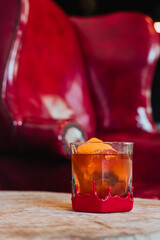 Amber cocktail in red-wax-dipped rock glass with ice sphere and orange-peel garnish set on wooden table against blurred red armchair background