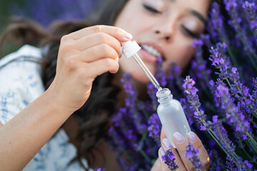 Closeup of a young beautiful dark haired woman applying serum to her face near lavender. Holding a...