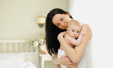 Happy young mother holding her cute baby standing together in white bedroom at home