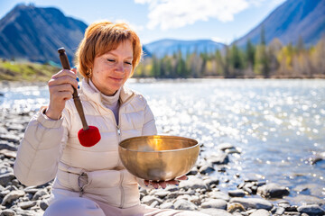 Woman performing a sound ritual with a Tibetan singing bowl by the Altai river. Concept of feminine...