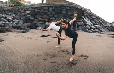 Two barefoot men practice advanced balance pose on sand, illustrating precision, muscular control...