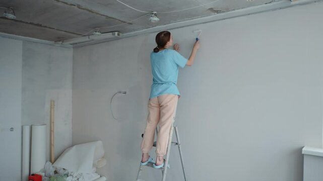 Woman painting upper wall with roller while standing on ladder