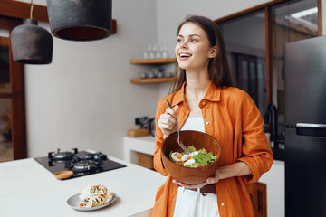 Healthy eating woman in an orange shirt joyfully holding a salad bowl in a modern kitchen setting,...