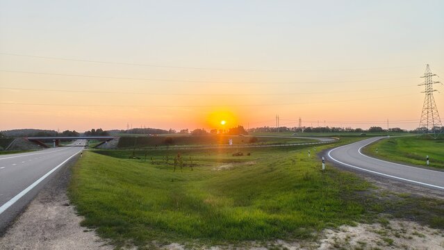 Next to the agricultural grassy meadow are asphalted roads and power lines on metal poles. Behind them, the summer sun sets and paints the sky and feather clouds in delicate colors - Powered by Adobe