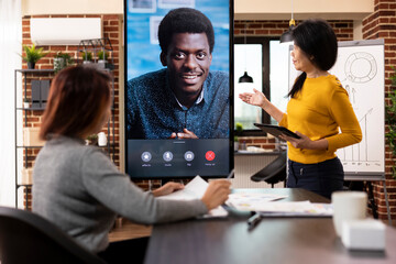 Female consultant stands with tablet, and coworker sits at desk, presenting company strategy ideas...